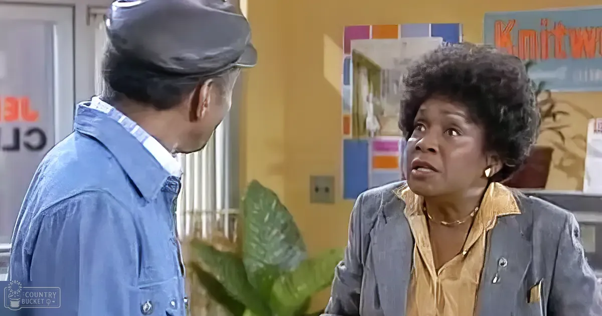 An adult female wearing a blue jacket with silver buttons and an adult male in a denim jacket interacting indoors, surrounded by plants and framed artwork, suggesting a casual office or community center setting during the daytime.