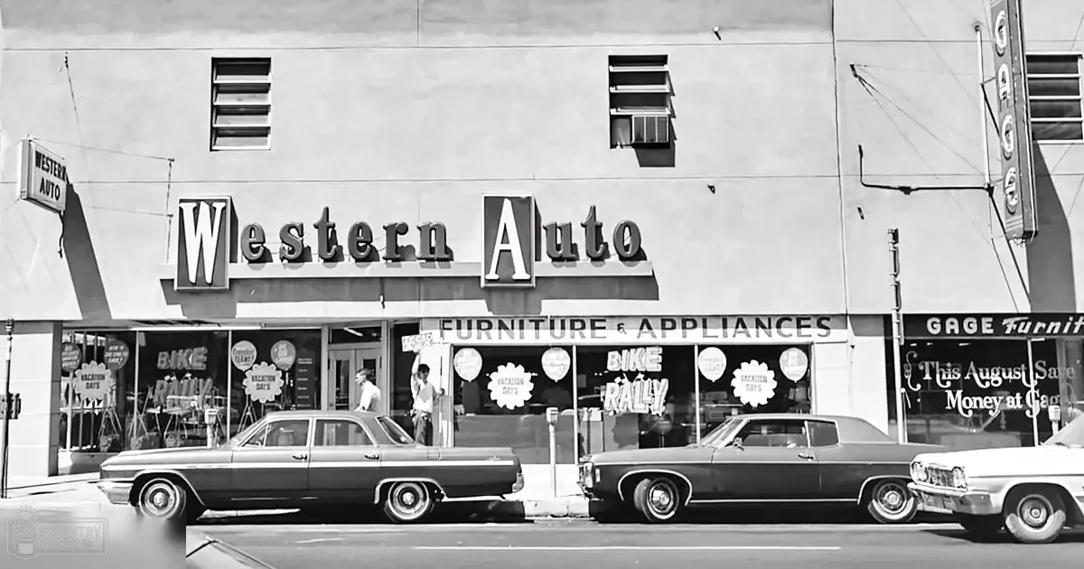 A Western Auto store interior with a young adult male in a white t-shirt and dark pants, another partially obscured individual in light-colored clothing, parked classic cars from the 1960s era, and signage for 'Gage Furniture' on an urban street during daytime summer or spring.