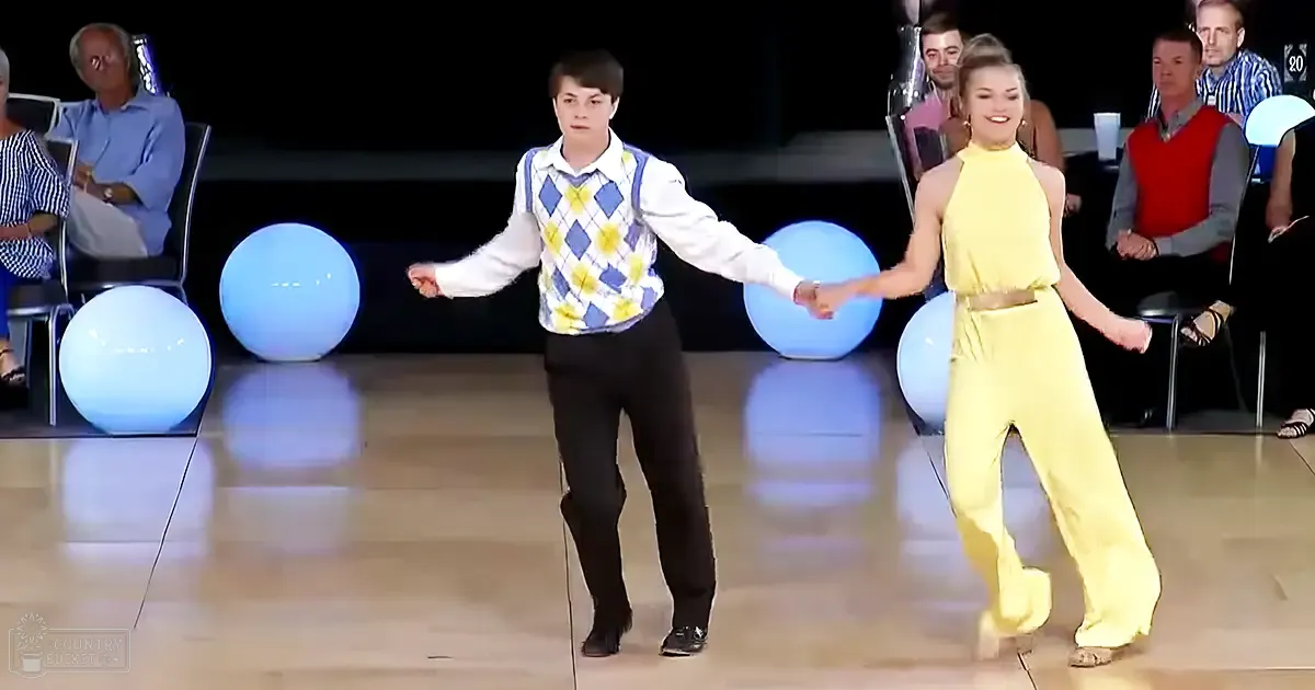 A dance pair performing on stage at an indoor event, with a young adult female wearing a bright yellow jumpsuit and a teenage male in a white shirt with blue and yellow argyle patterned vest over black trousers.