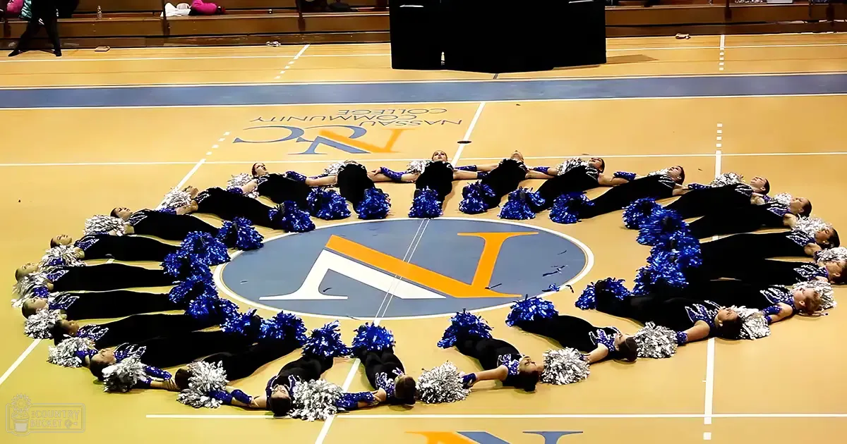 A cheerleading team performs in Nassau Community College gymnasium, with teenagers wearing black leotards adorned with silver and blue pom-poms. The setting is an indoor sports facility with bleachers and a well-lit environment.