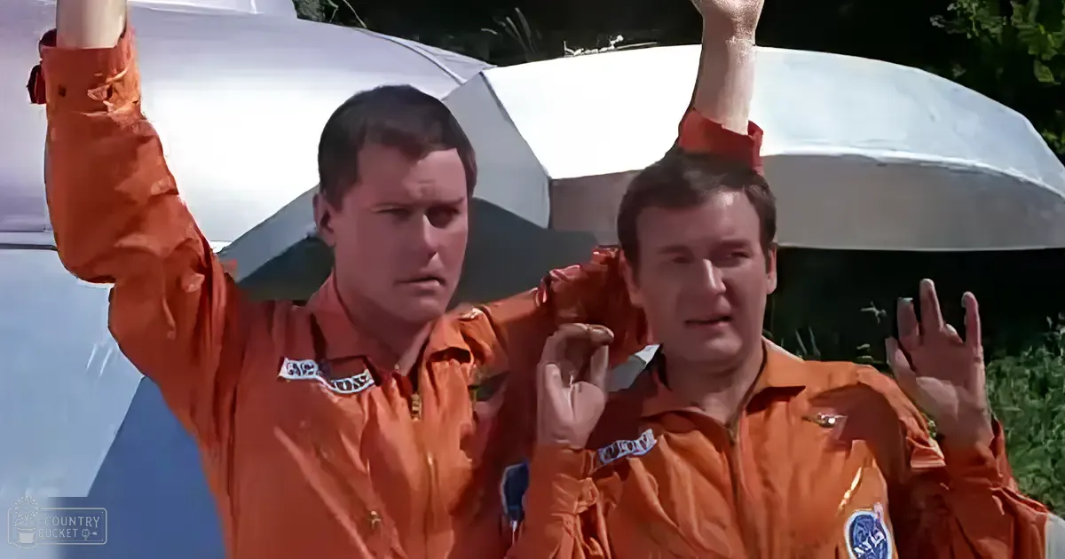 Two adults in orange jumpsuits stand outdoors, possibly on a film set with an aircraft canopy visible behind them.