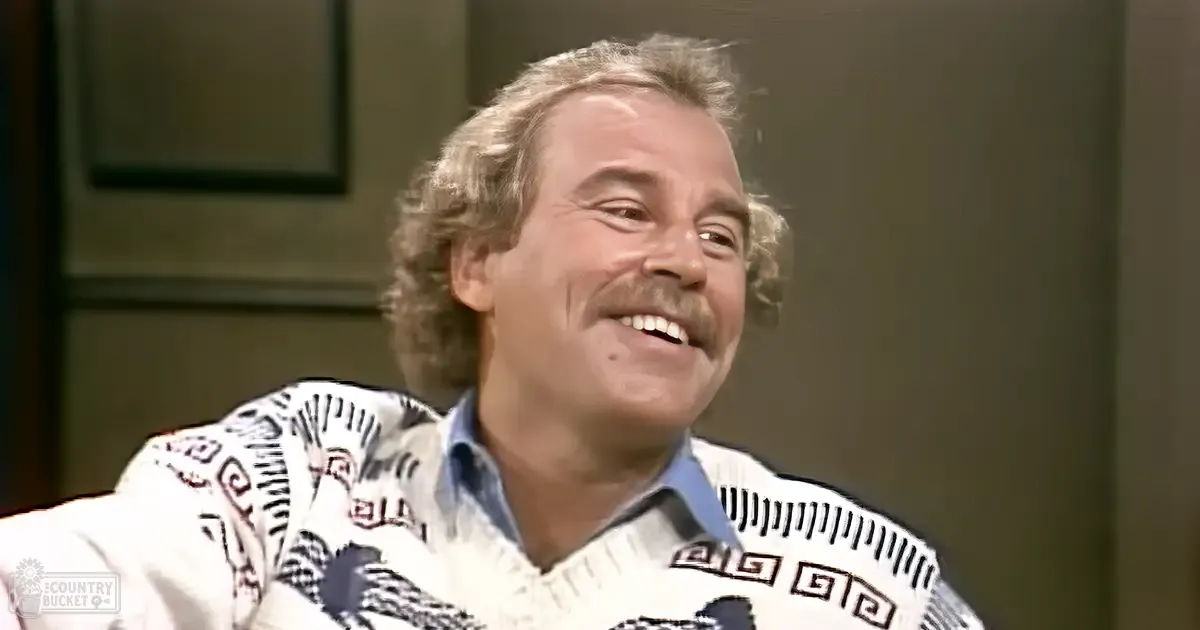 A middle-aged adult male wearing a patterned, relaxed-fit white sweater with black and red geometric designs and a collared shirt underneath. He appears to be in an indoor setting possibly for an interview or talk show.