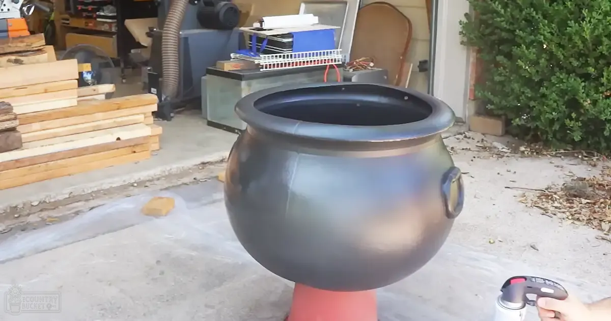 An outdoor workshop or garage with various tools, a large black pot on a red stand, a bush to the right, and scattered materials in the background.