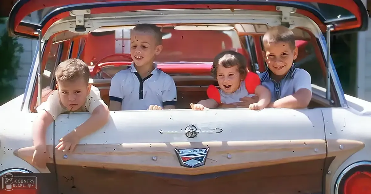 A group of children in a vintage Ford car outdoors, with white siding visible in the background.