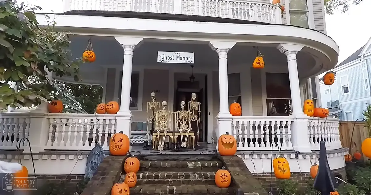 Halloween-themed Ghost Manor house with carved pumpkins, a 'Ghost Manor' sign, and skeleton figures in an urban residential area during late afternoon or early evening.
