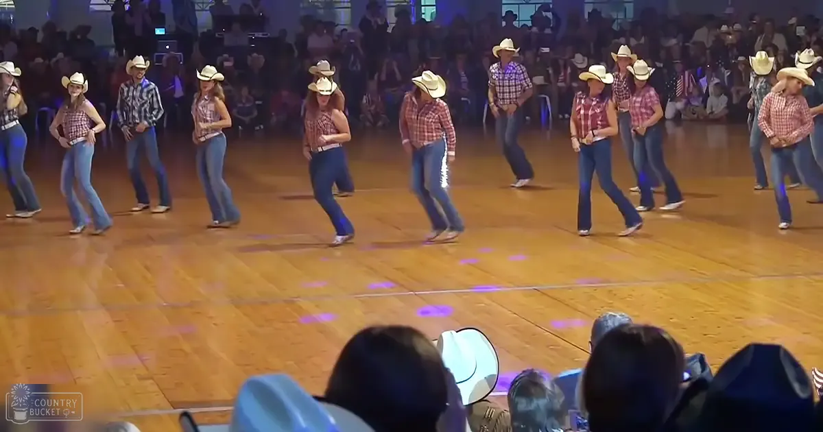 A group dance performance by young adult females in a casual indoor arena, wearing plaid shirts and cowboy hats under artificial evening lighting.