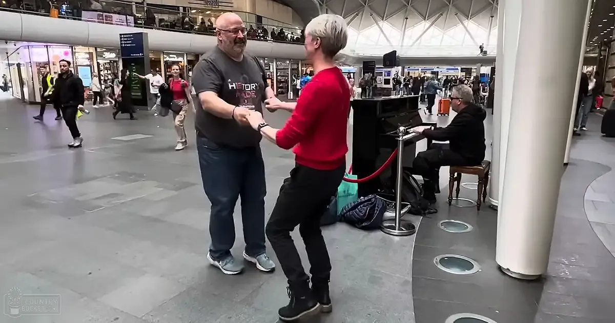 An adult male singer wearing glasses, a dark gray t-shirt with 'HISTORY' partially visible, blue jeans, light-colored sneakers, and an adult female partner in a red long-sleeve shirt, black pants, and dark boots dancing together in what appears to be an airport terminal. The setting includes various shops and people walking around, indicating an indoor public space like an airport or shopping mall.