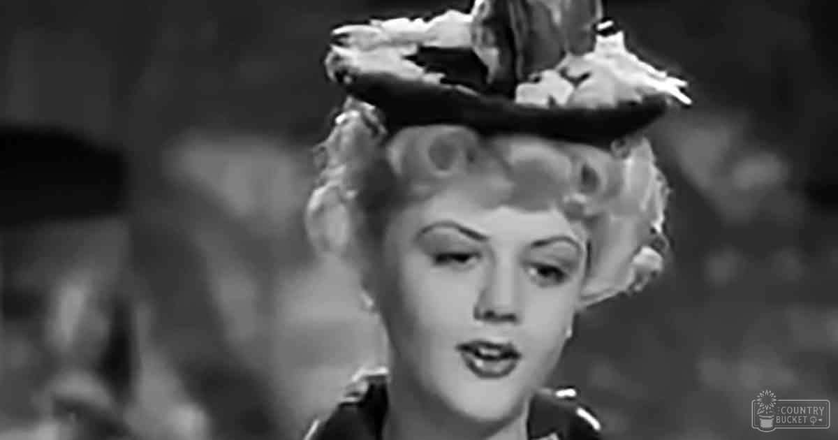 An adult female wearing a dark-colored garment with high collar, possibly mid-20th century, in an indoor setting with artificial lighting and blurred background shapes.