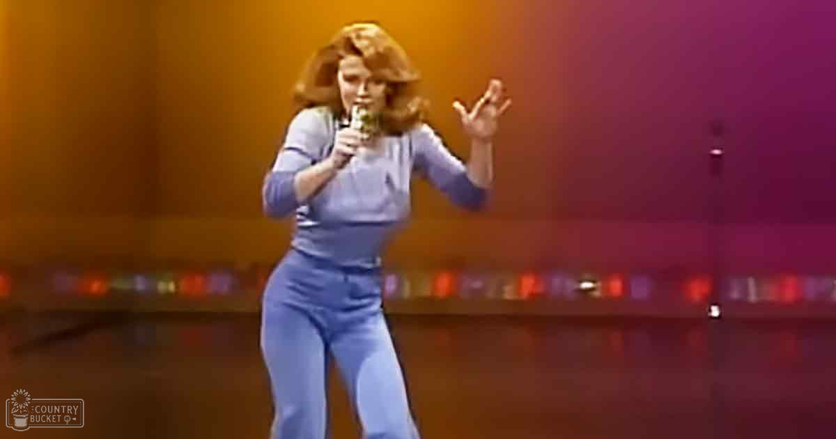 A middle-aged woman wearing a light blue 1970s-style jumpsuit sings into a microphone on stage with colorful lighting, suggesting a recording or performance studio setting.