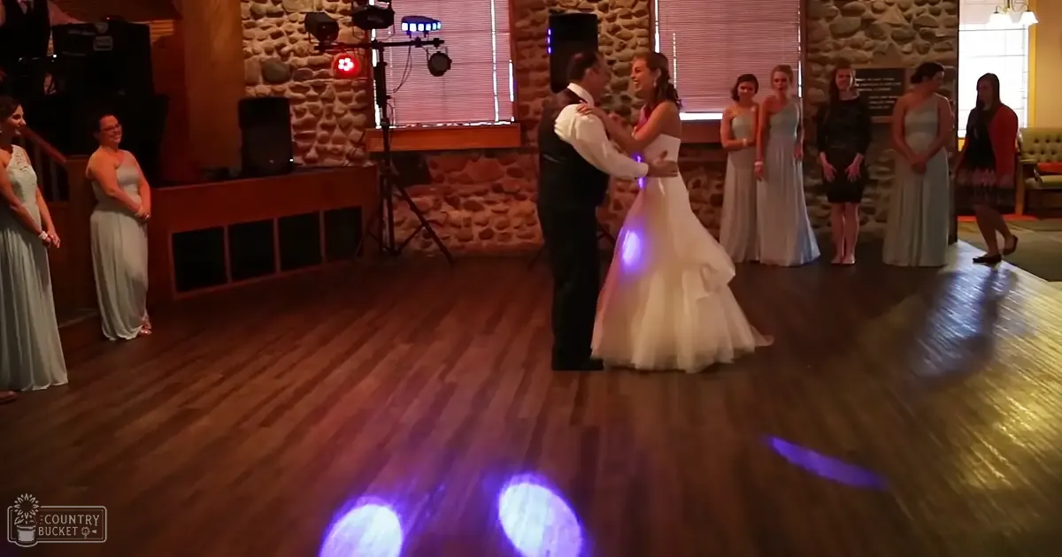 A rustic wedding reception scene with stage lighting and a couple dancing amidst bridesmaids and guests in an indoor venue at night.