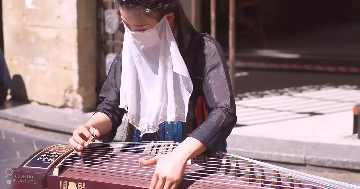 A young adult female plays a guzheng outdoors, wearing a black lace top and blue skirt, under bright sunlight with stone steps and scaffolding in the background.