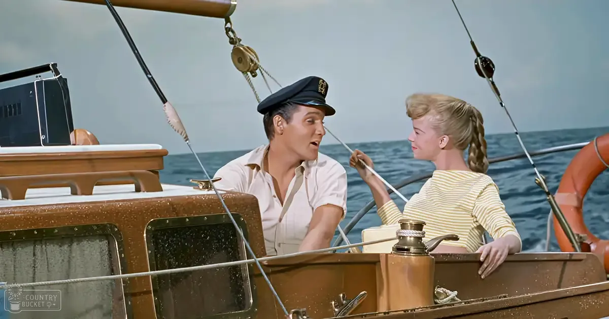 A maritime scene with a young adult male in a white shirt and captain's hat, alongside another young adult female wearing a yellow and white striped top. The setting is on a boat at sea under an overcast sky.