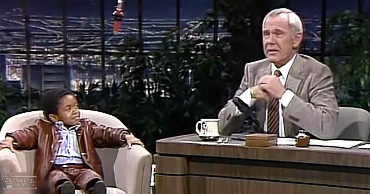A toddler male in a talk show set with an older adult male, both seated at a desk with a mug reading "The Tonight Show."
