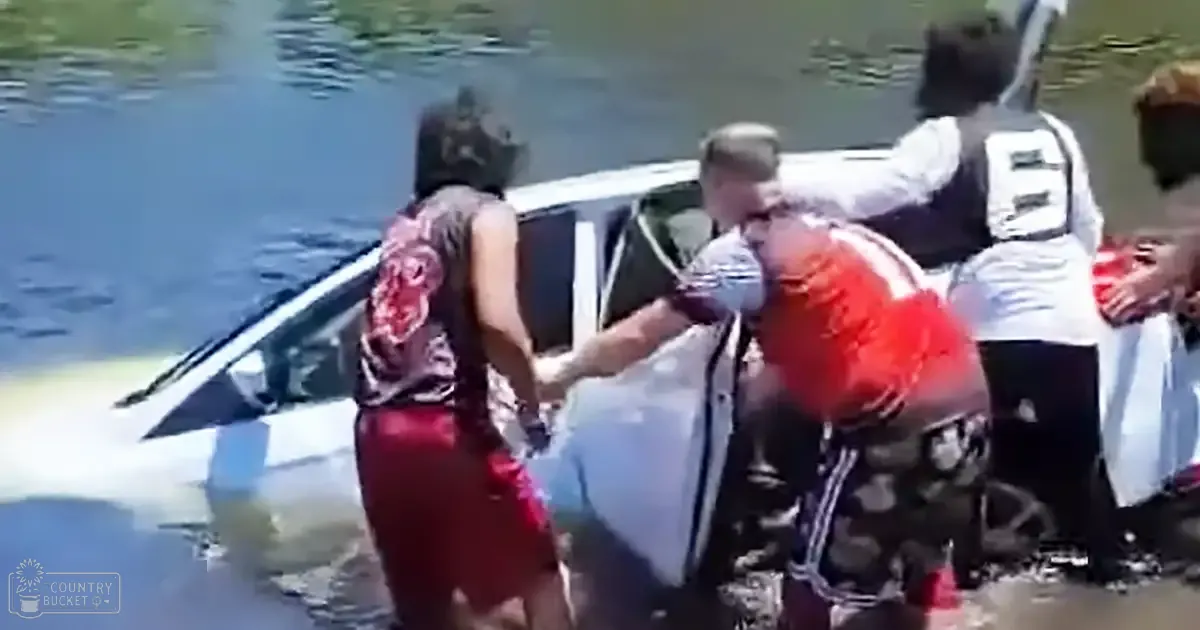 A rescue scene with several people near a partially submerged car in a body of water, likely after an accident; the setting is outdoors during daylight hours.
