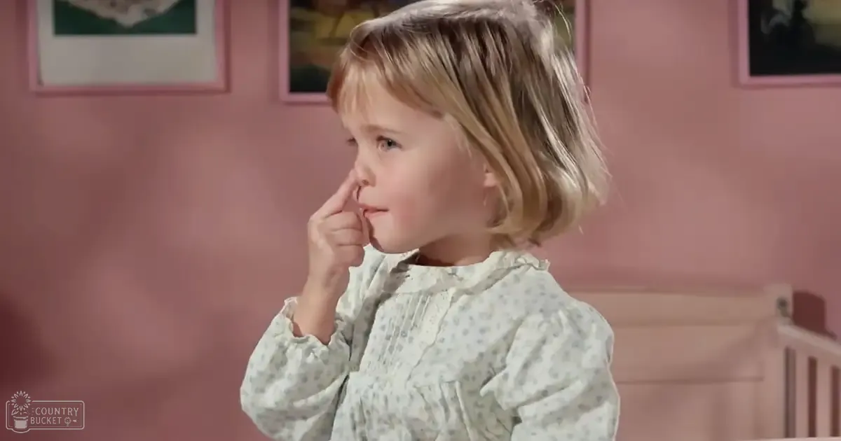 A toddler wearing a light green polka-dot dress with ruffled collar and long sleeves stands indoors near a crib in a softly lit bedroom, surrounded by pink-painted walls and framed artwork.