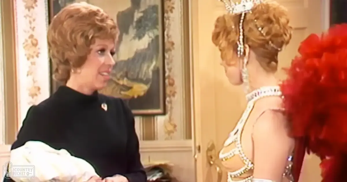An adult female in a black turtleneck sweater with white gloves, standing indoors with another woman wearing an elaborate white dress adorned with gold embellishments and a red feathered accessory. The setting appears to be a home or formal venue with natural lighting.
