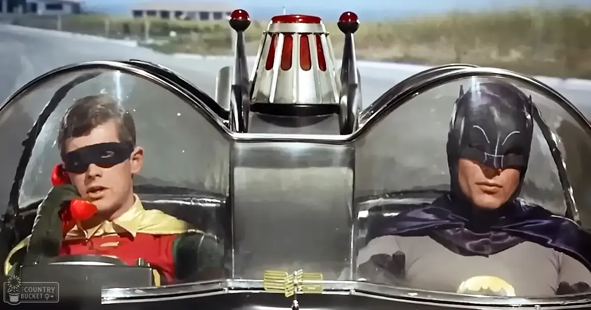 A scene from a classic superhero movie with two characters, one in a red and yellow costume with Robin's utility belt and cape, and another in Batman's dark gray suit with a bat emblem. The setting is an outdoor highway area during the daytime.