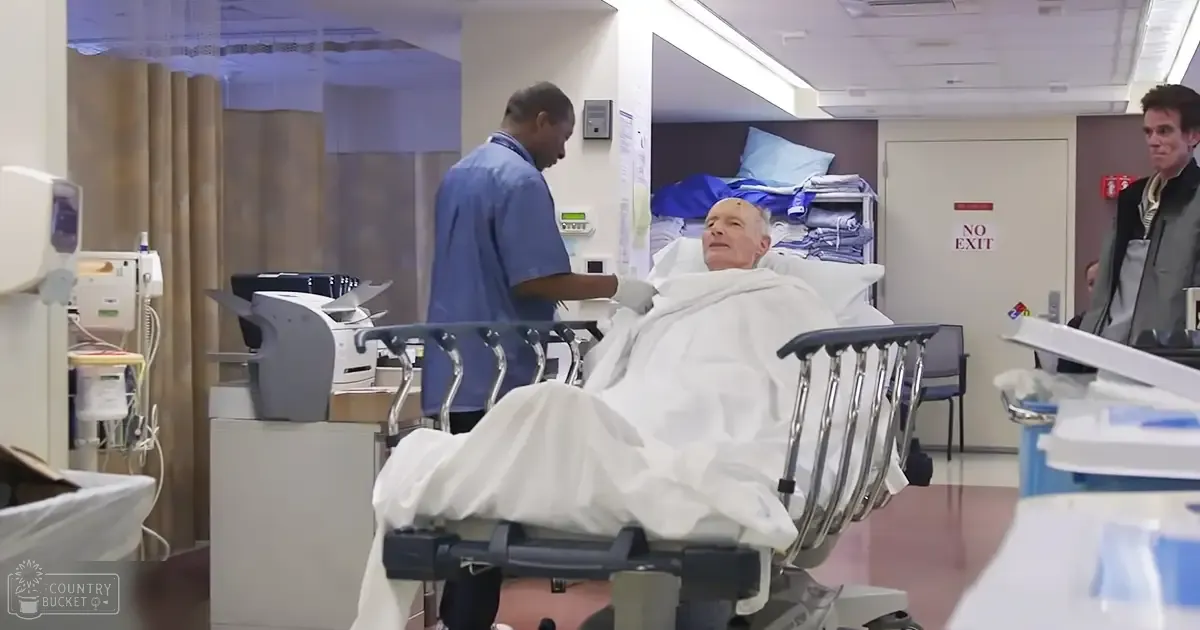 An older adult male patient lying in a hospital bed with white blankets, surrounded by medical staff and typical hospital furniture and equipment under artificial lighting.