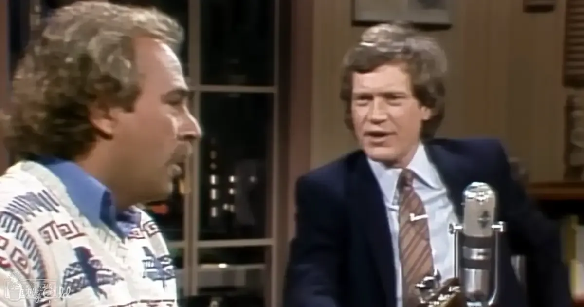 Talk show host and guest in a studio setting with late 20th-century attire, discussing under natural lighting conditions.