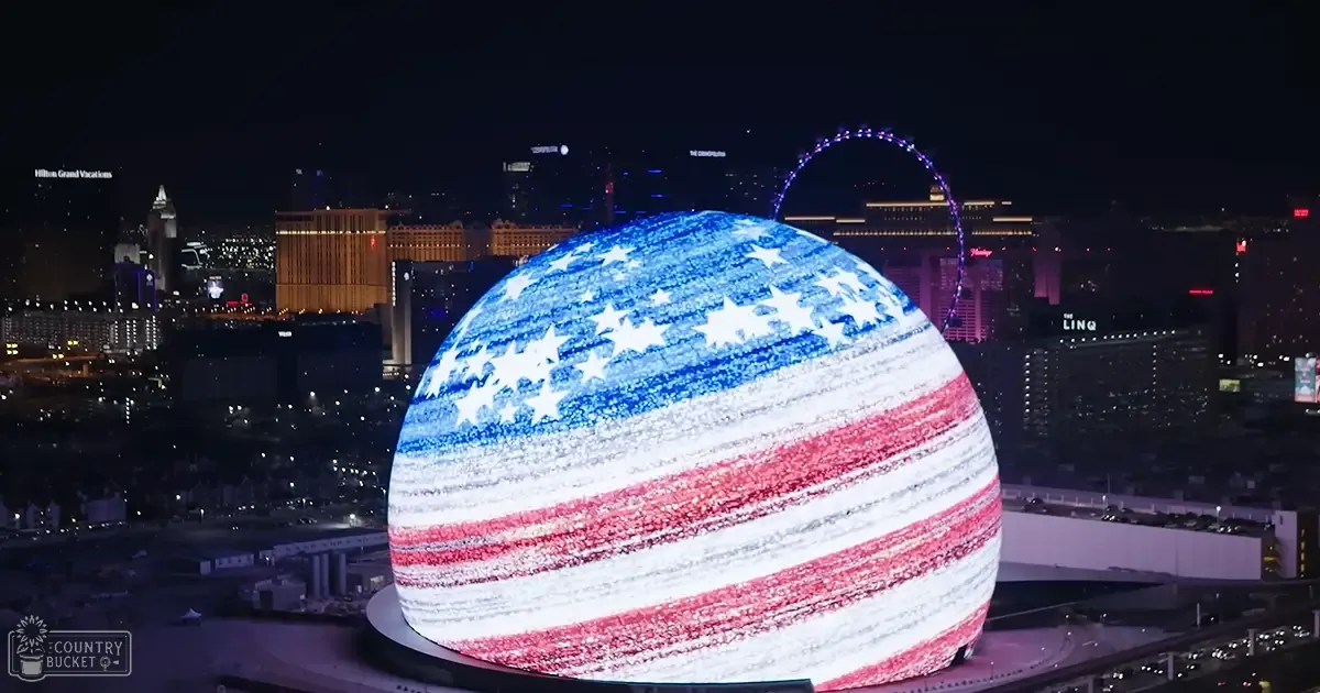 A nighttime view of Las Vegas with illuminated hotels and casinos, with a large spherical structure displaying an American flag motif against the city's skyline.