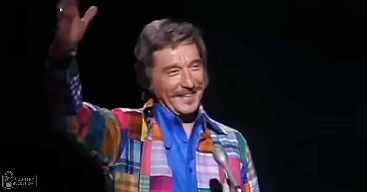 A middle-aged male performer in an indoor setting, wearing a plaid shirt with orange, blue, green, and white stripes. The scene takes place on a stage with dim lighting focused on the performer.