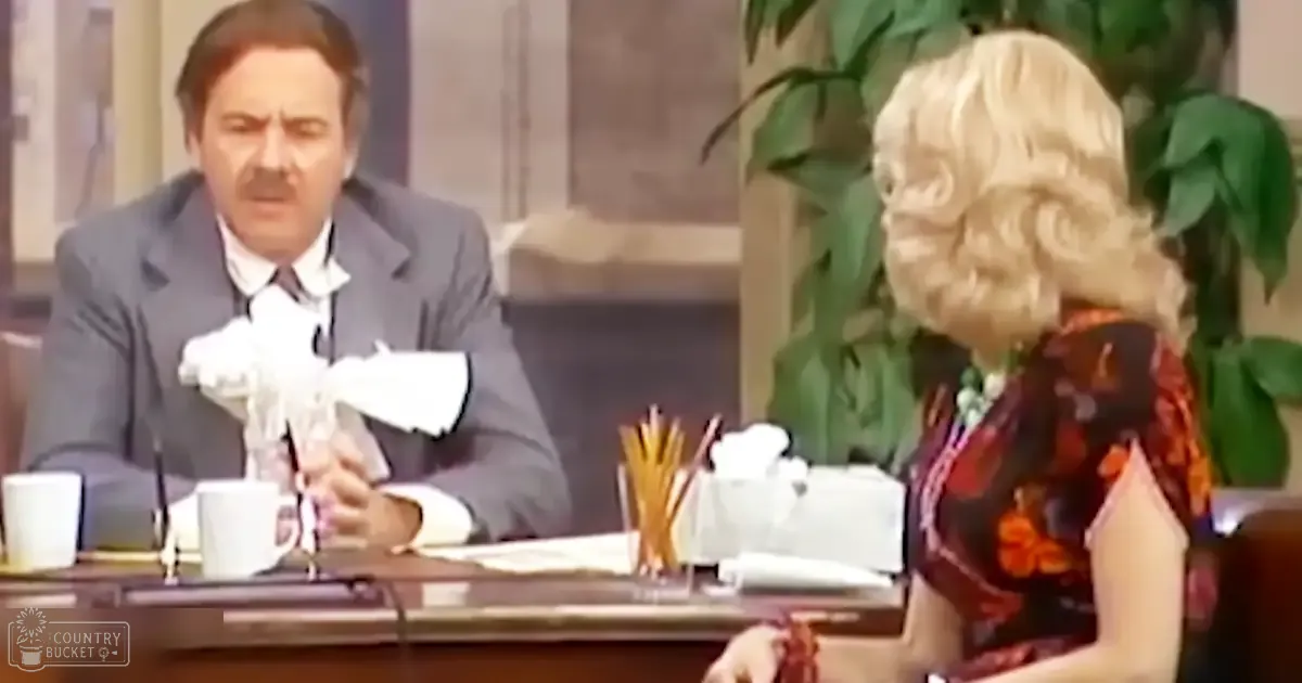 A middle-aged male and a woman in an office setting with detailed costumes, suggesting a theatrical or comedic scene.
