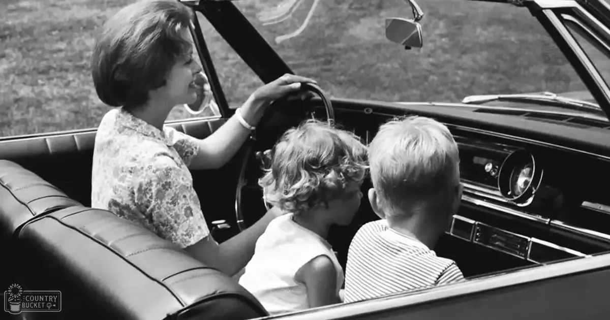 Middle-aged woman driving with two children in an open-top car on a grassy suburban area during what appears to be late morning or early afternoon.