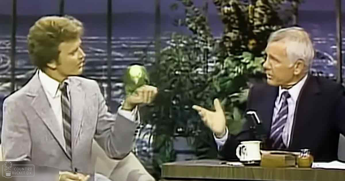 Two adult males in a talk show set, one wearing a light gray suit with diagonal-striped tie, the other in a dark suit and striped shirt, holding an Amazon parrot.