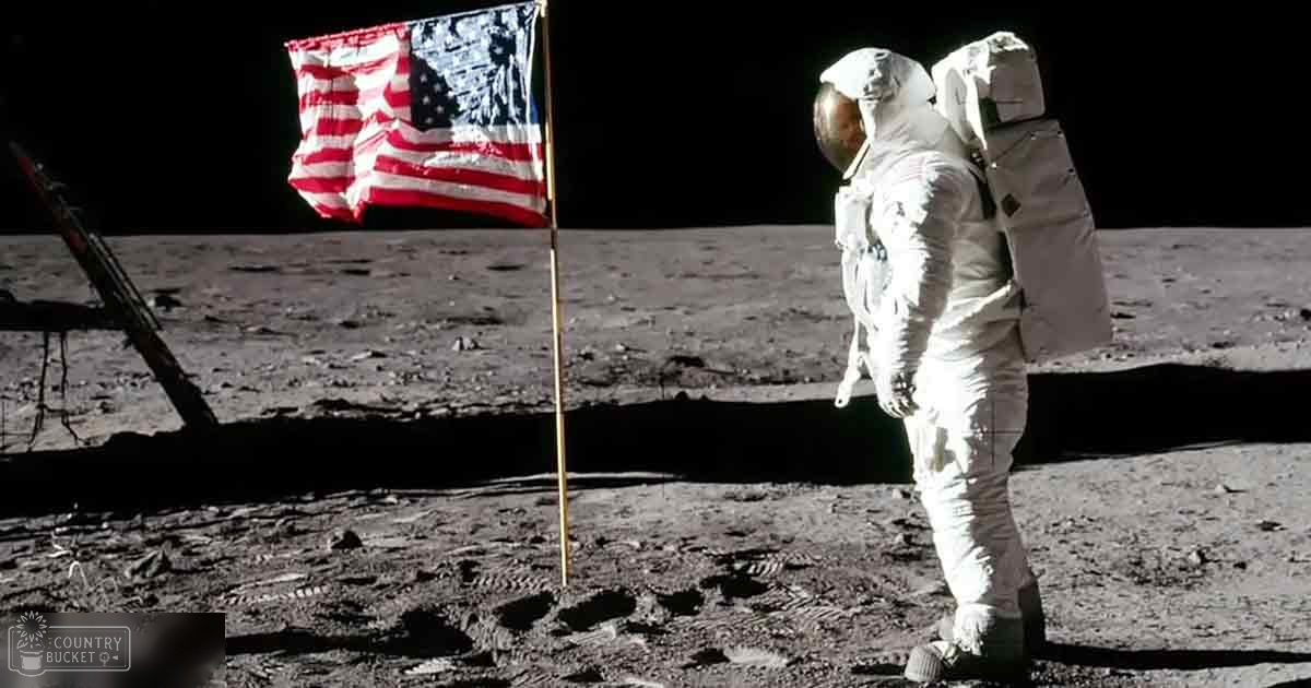 Astronaut in a white spacesuit stands next to an American flag on the moon's surface during daylight with clear visibility.