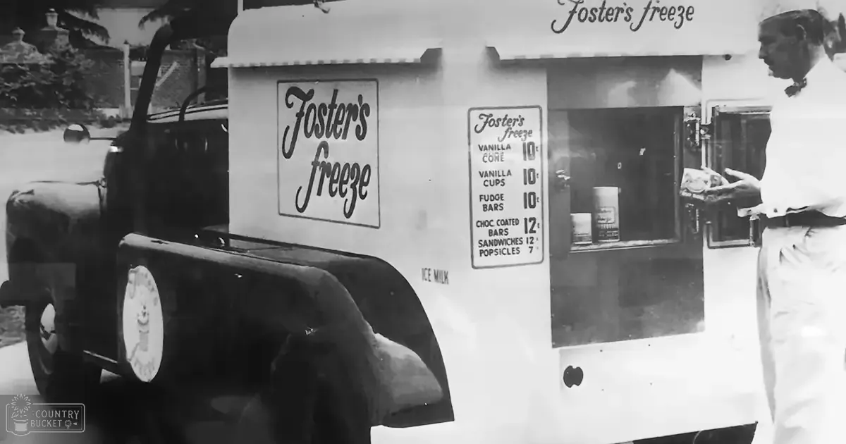 A vintage ice cream truck named 'Foster's Freeze' parked on a street in an urban setting, with part of the vehicle's rear and menu visible; a person stands beside it wearing a white shirt from the 1930s or 1940s.