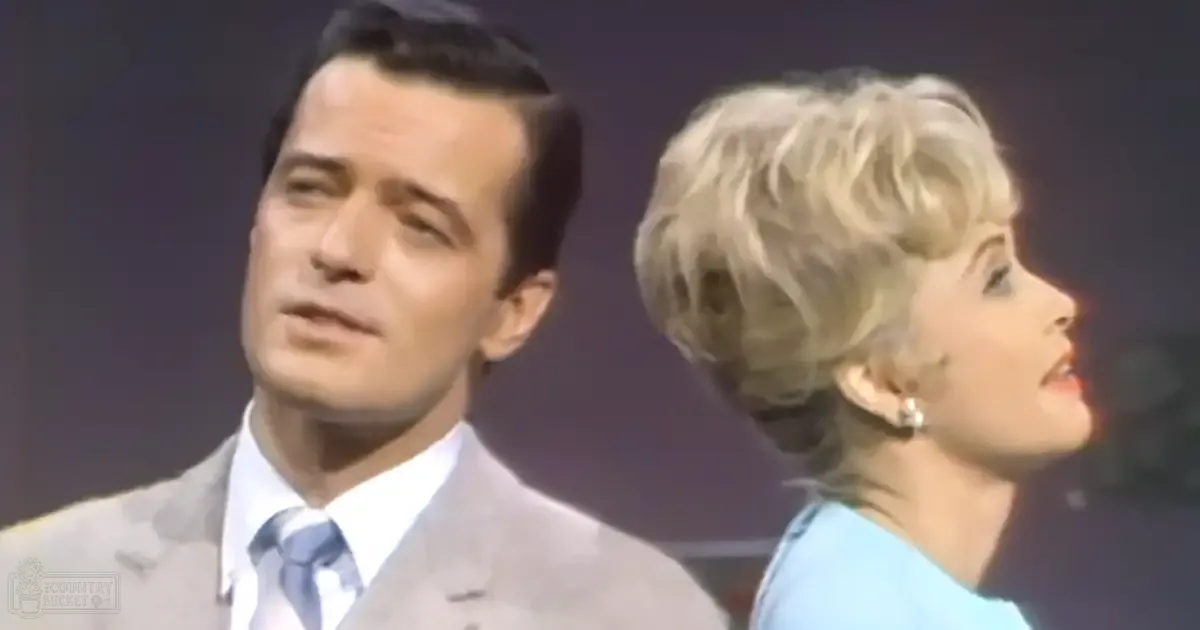 Adult male singer wearing a light gray suit and white dress shirt with a blue patterned tie, standing beside an adult female performer in a light blue top, possibly at an event or performance setting with dark backdrop indicating stage lighting.