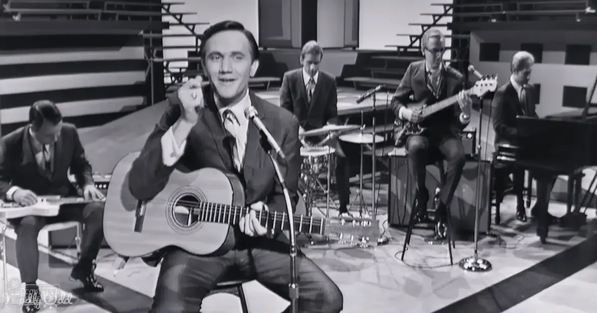 Adult male middle-aged musician playing guitar, surrounded by a band at an indoor recording studio or performance venue with musical instruments and microphones visible.