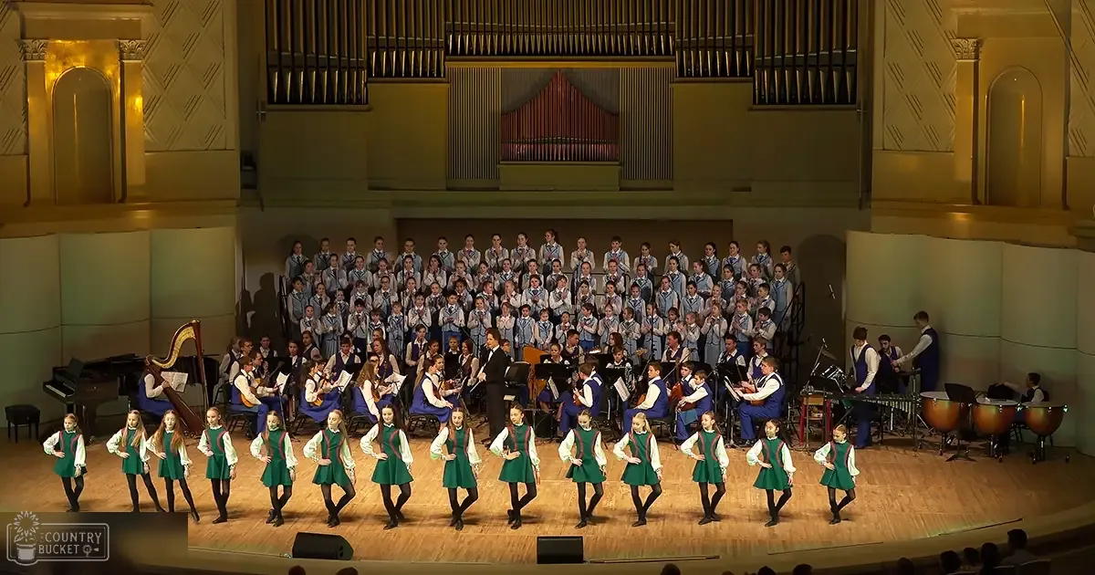 A choir and dancers perform in an ornate auditorium with wooden floors and organ pipes visible.
