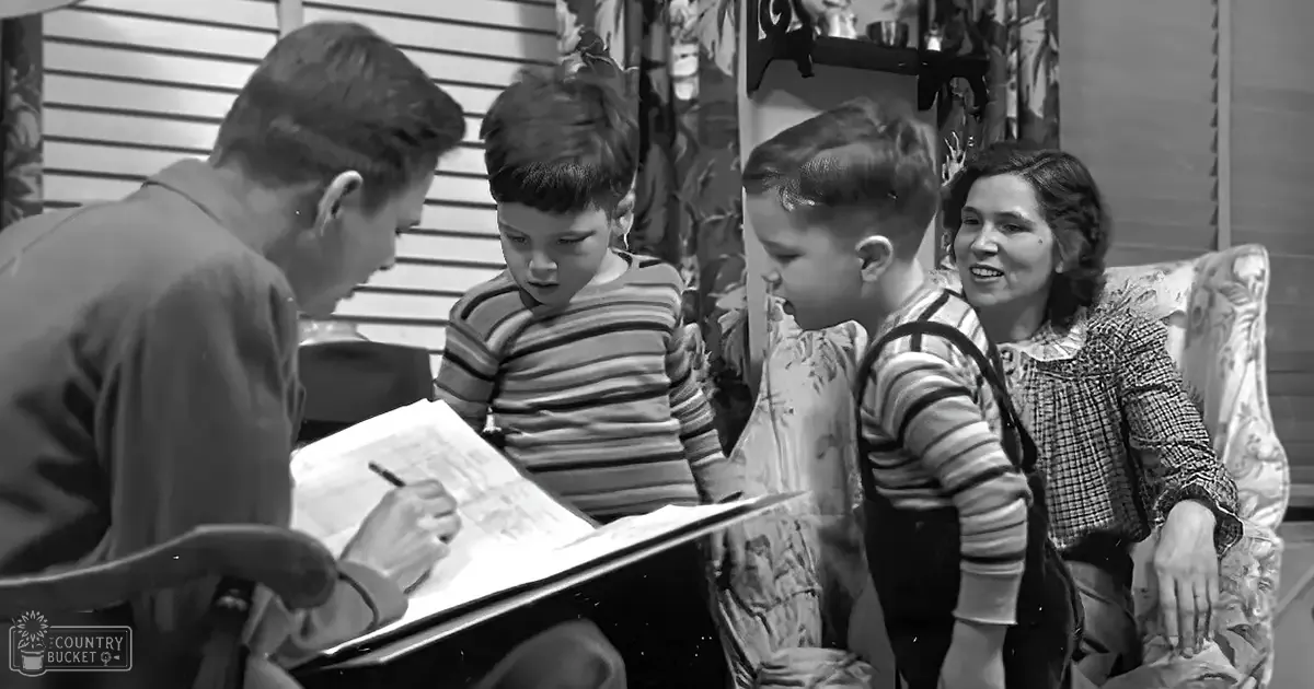 A young adult male seated at a table with two children around him in an indoor setting; the man holds a pen and wears a collared shirt, while one child is in striped long-sleeve overalls.