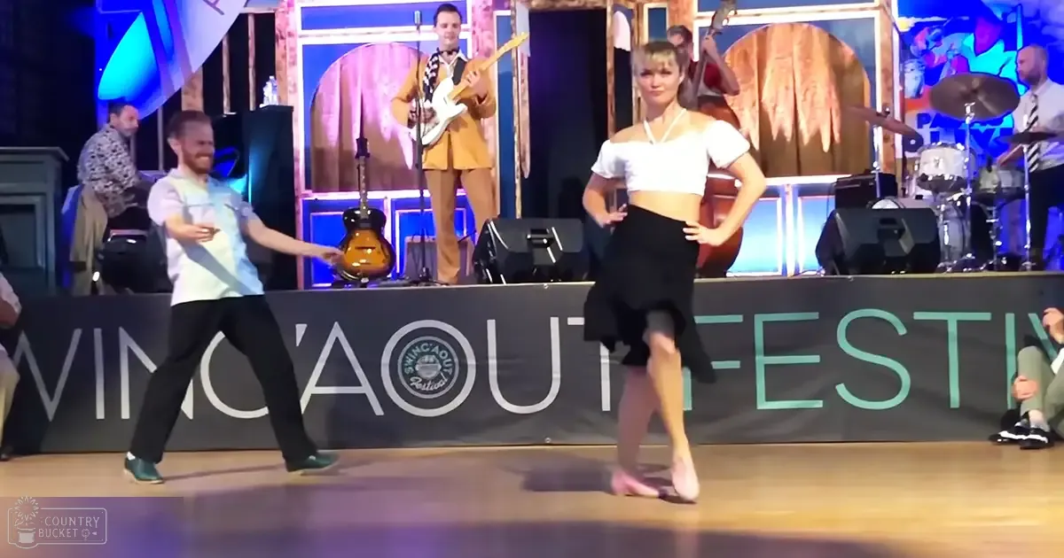 Dancers performing at the Shing'Aout Festival in a colorful indoor venue with stage lights and event banner.