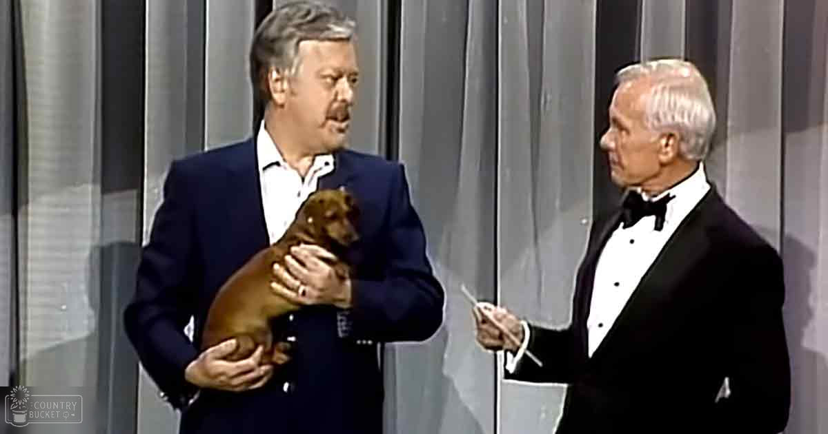 Two men in formal attire stand indoors, one holding a small Dachshund on stage under bright artificial lighting.