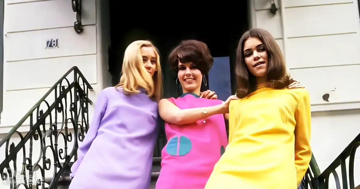 Young adults wearing dresses outdoors in front of a residential house with white siding and dark door frame, natural light suggesting daytime.