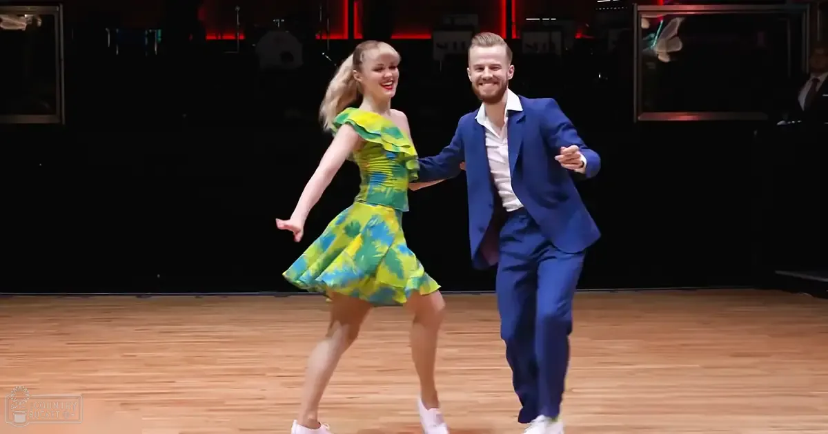 A young adult female dancer in a bright green floral dress and a male dancer in a blue suit jacket dance together in a warmly lit indoor studio setting.