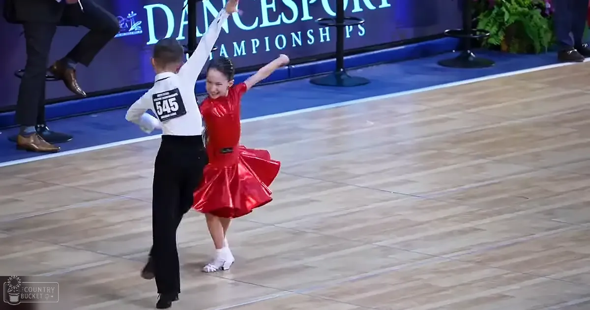 A young couple dances at a dance competition venue with wooden floors and plants near the stage.