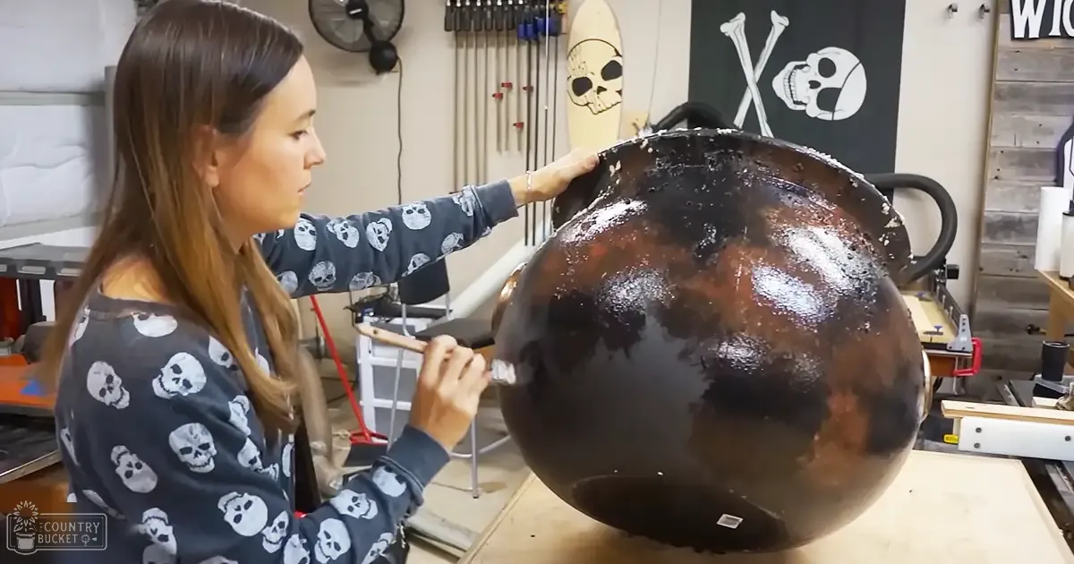 A young adult female in a workshop setting, wearing a dark gray or black long-sleeved sweatshirt with white skull patterns on the sleeves. Tools and equipment hang on the wall behind her, while a surfboard leans against it.