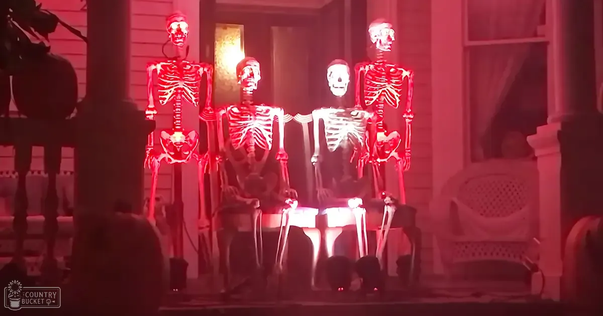 Four Halloween skeleton figures seated at a table in front of a house, with decorative elements like a horse statue to the left. The scene is illuminated by artificial light, suggesting nighttime or evening.