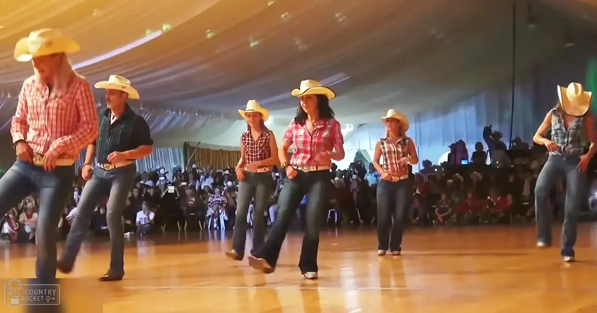 Group dance performance in an indoor setting with plaid shirts and cowboy hats, possibly vintage or retro style, under wooden flooring and tent-like ceiling structure.
