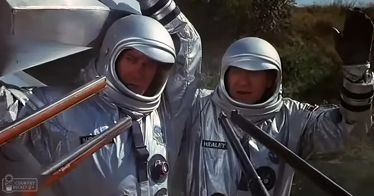 Two adult males in silver reflective space suits stand outdoors near a body of water, holding wooden poles; behind them is a large metallic structure suggesting a space-related environment.
