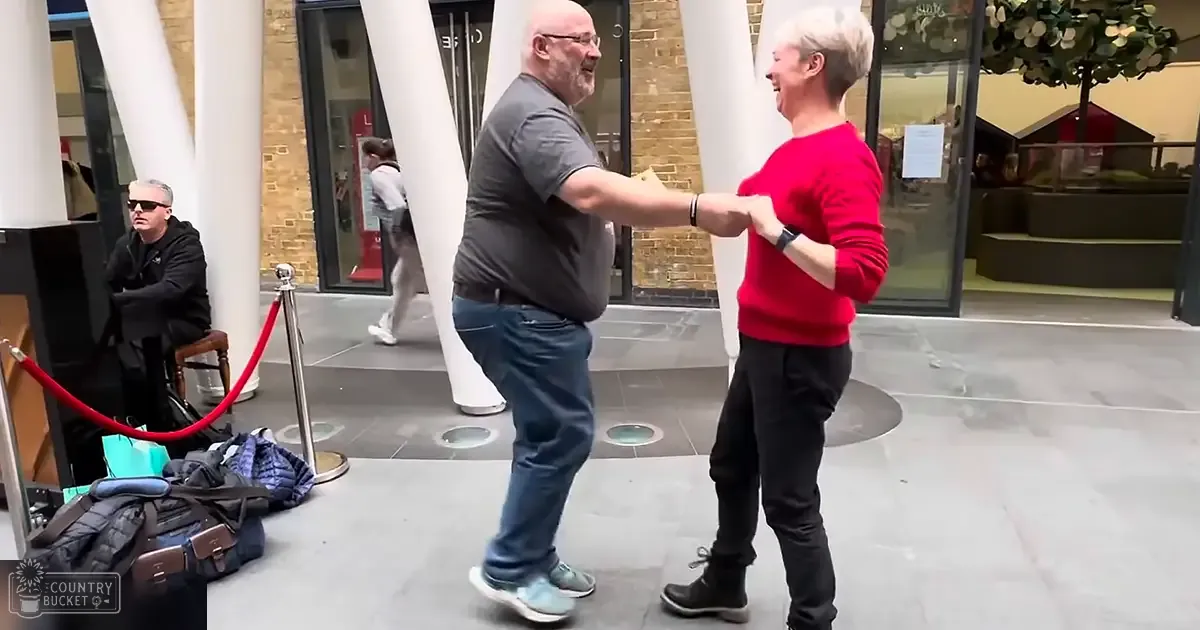 Two middle-aged adults dancing indoors; a man in dark gray T-shirt and jeans, woman in red sweater and black pants.