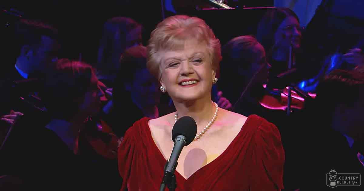 A middle-aged woman in a red puffed-sleeve dress performs on stage with musicians, audience visible.
