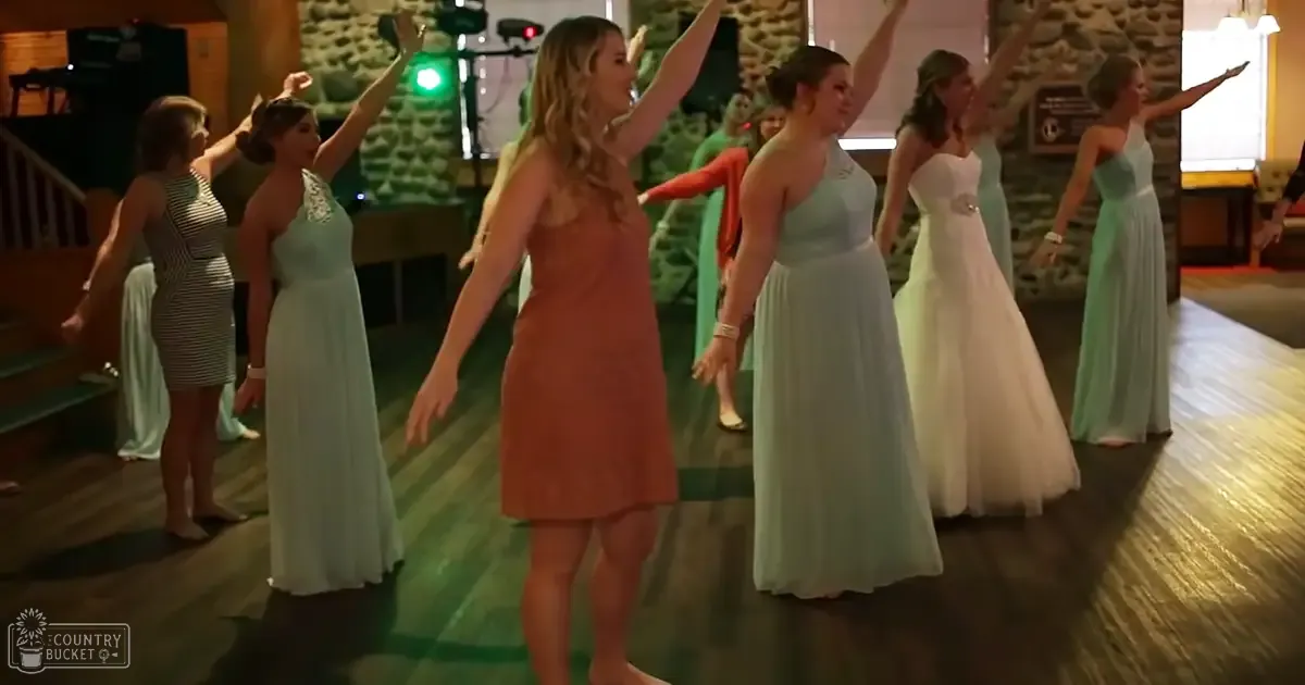 A group of women dancing in an indoor event space with wooden floors and stone walls under stage lighting.