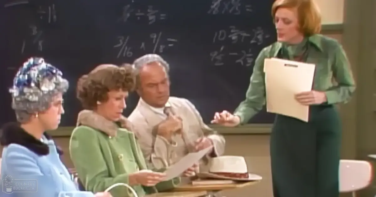 Middle-aged adult female teacher with short blonde hair and a green dress addresses students in a classroom setting, surrounded by desks, chairs, and educational furniture under even natural light.