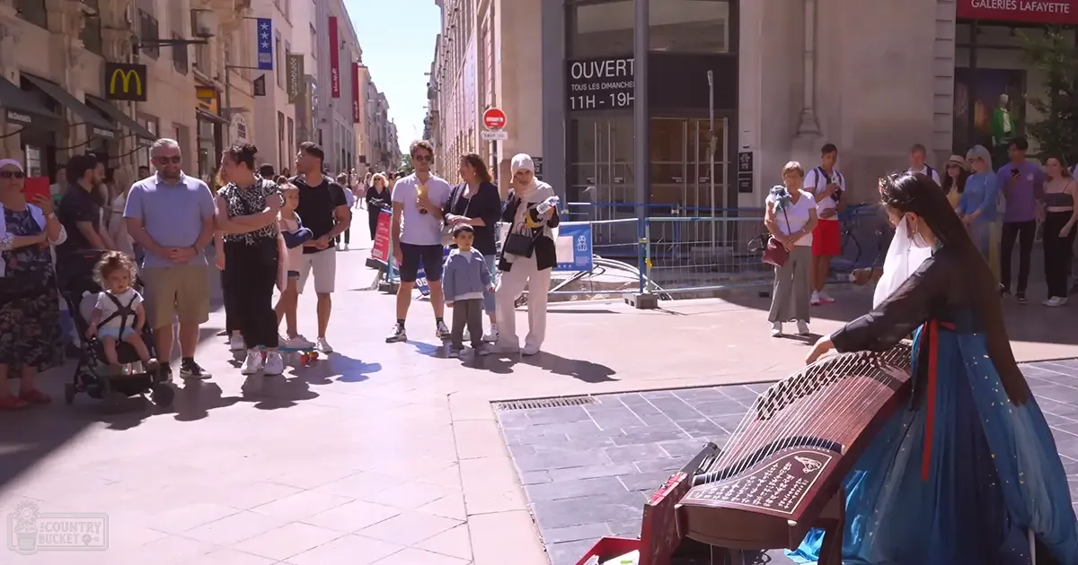 A street performer playing a zither in a sunny European city square with shops and people enjoying the day.