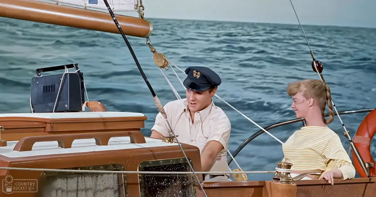 A man in his adult years wearing a white short-sleeved shirt and a dark blue captain's hat is on a boat with a young woman wearing a yellow and white striped top, her hair styled in pigtails. The setting shows an open body of water under clear skies, suggesting it might be summer or spring.