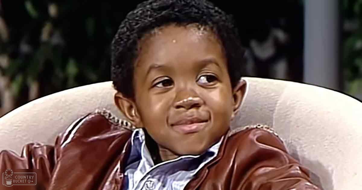 A toddler wearing a brown leather jacket with white trim and a blue-and-white striped collared shirt, seated indoors near a white armchair under soft, artificial lighting.